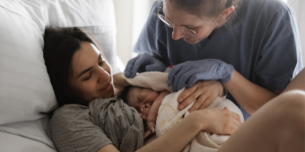 Hebamme freut sich mit der Mama über ein gesundes Neugeborenes und schaut wie es dem Kind geht. Das Kind liegt auf der Brust der Mutter und ist in ein Handtuch eingewickelt.