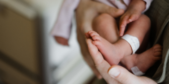 Frisch gebackene Mama hält die Babyfüße ihres Neugeborenen. Der Fokus liegt auf den Füßchen. Der Hintergrund ist unscharf.