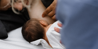 Nahaufnahme: Mama wird beim Stillen im Liegen begleitet. Das Baby trinkt an der Brust und Mutte und Stillberatern beobachten das Kind.