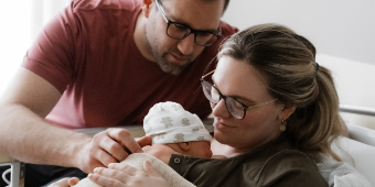 Frisch gebackene Eltern kuscheln mit ihrem Neugeborenen auf der Wochenbettstation. Die Mutter liegt mit leicht erhöhtem Oberkörper im Bett. Das Kind liegt bei ihr auf der Brust. Der Vater steht neben dem Bett und beugt sich zu den beiden. Mit einer Hand streichelt er dem Kind den Rücken.