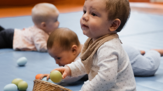 Nahaufnahme eines Kindes das im Kurs zu seiner Mama schaut. Es hält in der Hand ein Rasselei. Im Hintergrund krabbeln zwei andere Kinder über eine blaue Turnmatte.